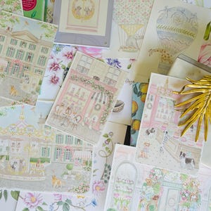 May include: A collection of illustrated art prints featuring whimsical scenes with dogs, including buildings, a carousel, and a hot air balloon. The prints have a pastel color palette with floral and architectural elements, and a gold leaf decoration.