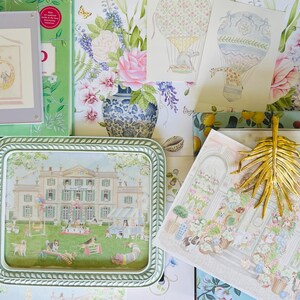 May include: A green and white ceramic tray with a picture of a house and a garden scene. The tray is surrounded by various decorative items, including a floral print, a book, and a gold palm leaf.