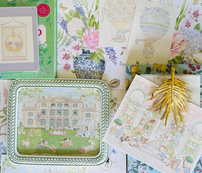 May include: A green and white ceramic tray with a picture of a house and a garden scene. The tray is surrounded by various decorative items, including a floral print, a book, and a gold palm leaf.