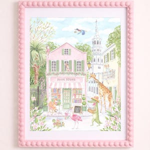 K&ouml;nnte beinhalten: Ein gerahmter Aquarelldruck einer skurrilen Buchladen-Szene. Das rosa Geb&auml;ude hat eine gestreifte Markise und die Worte "BOOK STORE". Eine Giraffe, ein Alligator und ein Flamingo geh&ouml;ren zu den Figuren. Der rosa Rahmen hat einen Perlenrand.