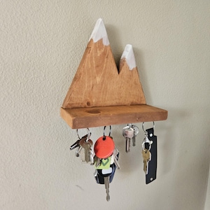 May include: Wooden mountain-shaped key holder with a shelf and several keys hanging from it. The mountain peaks are painted white. The key holder is mounted on a light-colored wall.