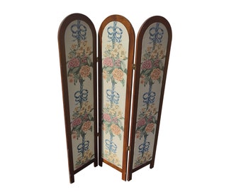 May include: A three-panel room divider with a floral and ribbon design. The panels are made of wood and have a dark brown finish. The fabric is a light beige with a floral pattern in shades of pink, blue, and green.