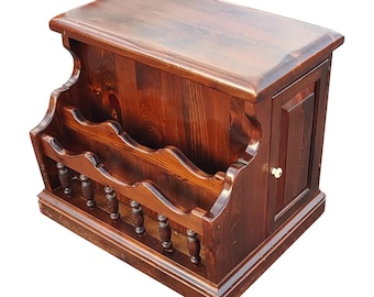 May include: A dark brown wooden magazine rack with a small cabinet door on the right side. The magazine rack has a decorative, scalloped design and a small shelf on the top.