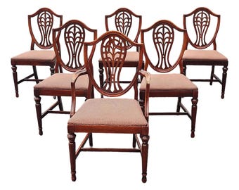 May include: Set of six antique wooden dining chairs. The chairs have a shield-shaped back with a carved design, upholstered seats, and curved legs. The wood is a rich, dark brown color, and the seats are a patterned brown fabric.