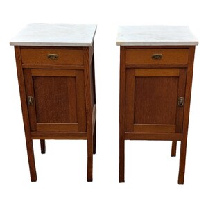 May include: A pair of wooden bedside tables featuring white marble tops, each equipped with a single upper drawer and a lower cabinet door. Both units include brass hardware pulls on the drawer and door, supported by four straight wooden legs.