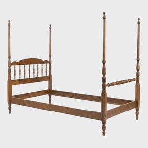 May include: A wooden four-poster bed frame with a headboard featuring vertical spindles and a curved top. The bed frame is made of a light brown wood and has four tall posts at each corner. The word "GAIL" is engraved on the side of the bed frame.