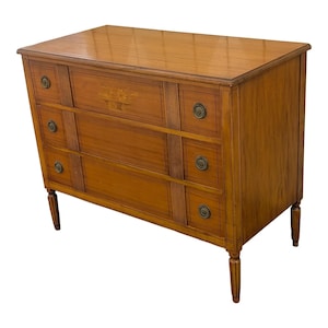 May include: A wooden chest of drawers with three drawers and brass knobs. The chest is a warm brown color with a glossy finish. The top drawer has a floral inlay. The chest has four slender legs.