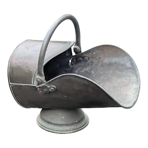 Può includere: Secchio per carbone in metallo antico con manico curvo e parte anteriore a forma di paletta. Il secchio è montato su una base rotonda. Il metallo ha un aspetto ossidato e invecchiato, che suggerisce che sia vintage. Il design è funzionale e decorativo.