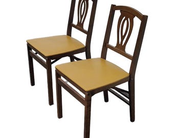 May include: Two wooden chairs with a dark brown frame and a light brown seat. The chair backs have a decorative oval design. The chairs are set against a white background.