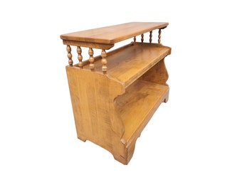 May include: A wooden shelf unit with two shelves and a top surface. The shelf unit has a decorative spindle design on the front and is made of light brown wood.