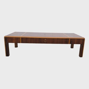 May include: A rectangular coffee table crafted from dark brown wood with a prominent grain pattern. The table features a long, horizontal drawer across the front, accented by a circular metal pull handle centered on the drawer face.