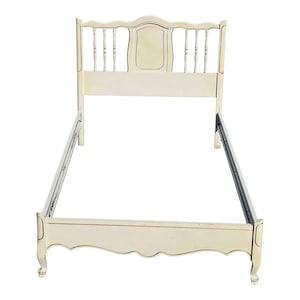 May include: Cream-colored bed frame with a decorative headboard. The headboard features a central panel and vertical spindles. The frame has a curved footboard and ornate legs. The bed frame is made of wood and has a distressed finish.