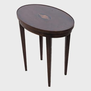 May include: Oval-shaped, dark brown wooden side table with four slender legs. The tabletop features a decorative inlaid design in the center. The table is approximately 28 inches tall and 20 inches wide.