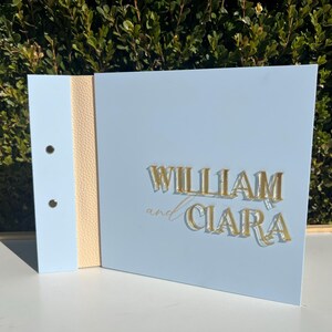 May include: A light blue wedding guest book with a beige ribbon down the spine. The cover has the names "William and Ciara" in gold lettering.