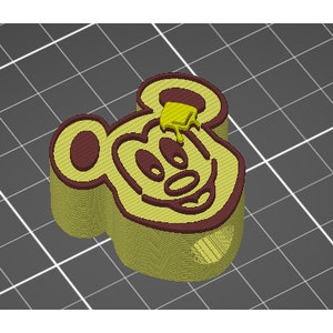 Op de afbeelding: Een gele 3D-geprinte Mickey Mouse-kop met een bruine omtrek. De kop staat op een grijze rasterachtergrond.