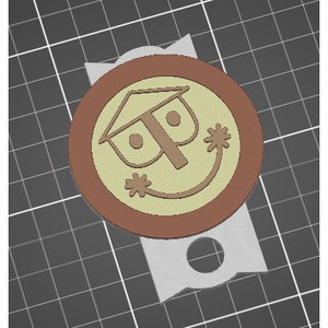 Puede incluir: Un emblema circular marrón y beige impreso en 3D con una cara sonriente con cabeza en forma de casa y manos en forma de estrella. El emblema está unido a una base gris claro en forma de cinta. El diseño recuerda a un icono clásico de parque temático.