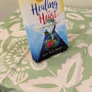 K&ouml;nnte beinhalten: Ein Buch mit dem Titel "Healing of the Heart" mit einem farbenfrohen Cover, das eine Sonne und abstrakte Elemente zeigt. Das Buch liegt auf einer gr&uuml;nen Tischdecke mit wei&szlig;em Blumenmuster.