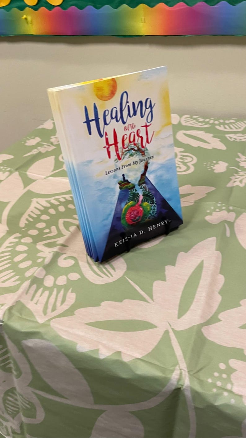 K&ouml;nnte beinhalten: Ein Buch mit dem Titel "Healing of the Heart" mit einem farbenfrohen Cover, das eine Sonne und abstrakte Elemente zeigt. Das Buch liegt auf einer gr&uuml;nen Tischdecke mit wei&szlig;em Blumenmuster.