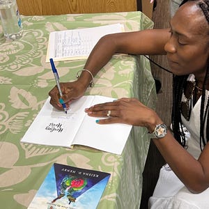 K&ouml;nnte beinhalten: Eine Frau signiert ein Exemplar von "Healing of the Heart" von Keisha D. Henry. Das Buchcover zeigt ein farbenfrohes Design mit dem Titel in einer stilisierten Schriftart. Ein Stift und ein H&auml;ndedesinfektionsmittel liegen auf dem Tisch, der mit einer gr&uuml;n-wei&szlig; gemusterten Tischdecke bedeckt ist.