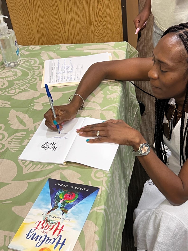 K&ouml;nnte beinhalten: Eine Frau signiert ein Exemplar von "Healing of the Heart" von Keisha D. Henry. Das Buchcover zeigt ein farbenfrohes Design mit dem Titel in einer stilisierten Schriftart. Ein Stift und ein H&auml;ndedesinfektionsmittel liegen auf dem Tisch, der mit einer gr&uuml;n-wei&szlig; gemusterten Tischdecke bedeckt ist.