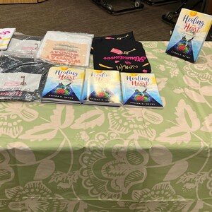 K&ouml;nnte beinhalten: Ein Tisch mit einem floral gemusterten Stoff, auf dem B&uuml;cher und Kleidung ausgestellt sind. Das Buch "Healing Heart" ist zu sehen, zusammen mit verpackten T-Shirts und einem schwarzen Shirt mit Text und Grafiken. Eine rote Papiert&uuml;te ist ebenfalls vorhanden.