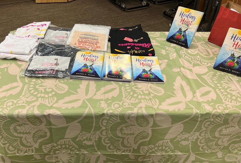 K&ouml;nnte beinhalten: Ein Tisch mit einem floral gemusterten Stoff, auf dem B&uuml;cher und Kleidung ausgestellt sind. Das Buch "Healing Heart" ist zu sehen, zusammen mit verpackten T-Shirts und einem schwarzen Shirt mit Text und Grafiken. Eine rote Papiert&uuml;te ist ebenfalls vorhanden.