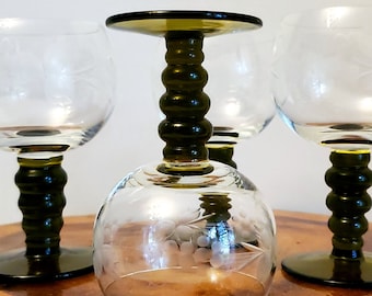 May include: Four clear wine glasses with etched floral patterns. The stems are dark green with a bulbous, ridged design. One glass is inverted, resting on a wooden surface.