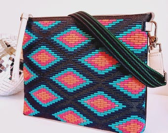 May include: A rectangular crossbody bag with a woven geometric pattern in shades of black, teal, orange, and pink. The bag has a black and green striped strap, a white wrist strap, and silver hardware.
