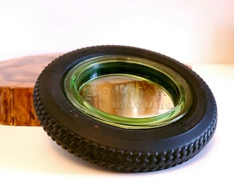 May include: A retro ashtray with a black tyre rim and a green glass interior. The glass insert is etched with the words "Goodrich Silvertowns". The ashtray is displayed on a white surface, with a wooden slice in the background.