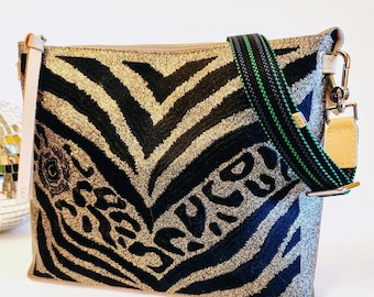 May include: A rectangular crossbody bag with a gold and black zebra print design. The bag has a black and green strap, a beige leather strap, and a zip closure. The word "ROCK" is printed on the front of the bag.
