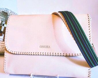 Puede incluir: Un bolso bandolera de cuero rosa claro con cierre de solapa. El bolso tiene una correa a rayas en verde, azul y negro. La palabra "CONSUELA" está impresa en la parte delantera. Los bordes están cosidos con un hilo en contraste.
