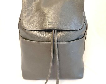 May include: Grey leather backpack with a flap closure and drawstring. Features two front pockets and a top handle. The brand name "CONSUNA" is subtly embossed on the flap. The bag is made of a textured leather.