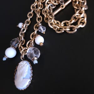 Puede incluir: Un collar vintage dorado con un colgante de camafeo y cuentas colgantes. El camafeo ovalado presenta un perfil en blanco sobre un fondo azul claro. El collar incluye cuentas blancas y transparentes.