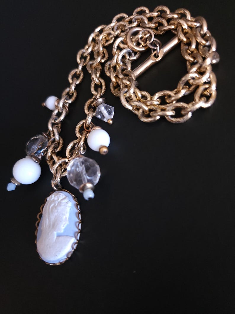 Puede incluir: Un collar vintage dorado con un colgante de camafeo y cuentas colgantes. El camafeo ovalado presenta un perfil en blanco sobre un fondo azul claro. El collar incluye cuentas blancas y transparentes.