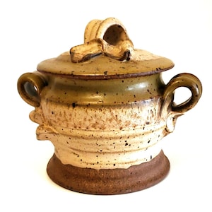 May include: A handmade ceramic sugar bowl with a lid and handles. The bowl is a mix of beige, brown, and green hues, with a textured surface and speckled details. The lid features a decorative handle. A rustic, handcrafted kitchenware item.