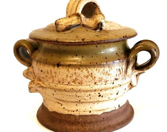 May include: A ceramic sugar bowl with a lid and handles. The bowl is a mix of beige, brown, and green hues, with a textured surface and speckled details. The lid features a decorative handle. A rustic, handcrafted kitchenware item.