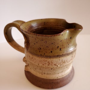 May include: Handmade ceramic pitcher with a handle. The pitcher features a speckled, earthy design with a brown base, a tan midsection, and a green-brown top. The pitcher has a spout for easy pouring and is ideal for serving beverages.