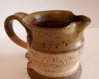 May include: Handmade ceramic pitcher with a handle. The pitcher features a speckled, earthy design with a brown base, a tan midsection, and a green-brown top. The pitcher has a spout for easy pouring and is ideal for serving beverages.