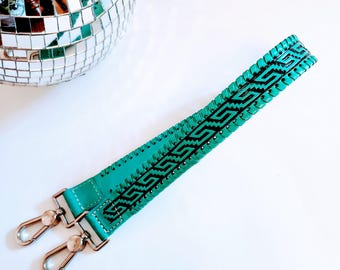 May include: A turquoise leather bag strap with a black geometric design. The strap has silver-coloured metal clasps on each end. The strap is approximately 102 cm long.