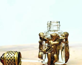 May include: A clear glass perfume bottle with a gold-coloured cherub design. The bottle has a decorative stopper with a red gem. The bottle is on a reflective surface.