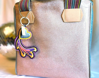 May include: A pink glitter tote bag with a colourful striped handle and a decorative charm. The bag has a light brown handle and trim, with a blue accent on the side. The charm is heart-shaped with a colourful, stylised design.