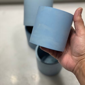 May include: Two light blue ceramic planters, one held in a hand, the other on a table. The planters are cylindrical and have a smooth, matte finish.