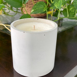 May include: A white concrete candle holder with a white candle inside. The candle is lit and has a wick.