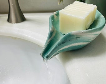 May include: A green and white ceramic soap dish with a spout, holding a white bar of soap. Water is flowing from the spout into a white sink.