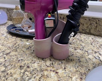 May include: A pink hair dryer and a black hair straightener are stored in a light pink ceramic holder on a bathroom countertop. The hair dryer is wrapped with a black cord. The countertop has a speckled, tan and brown pattern.