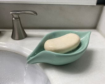 May include: A light green, leaf-shaped soap dish holds a bar of soap. The soap dish is made of ceramic and sits on a white countertop next to a silver faucet. Water is running from the faucet into the sink.