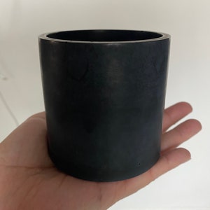 May include: A cylindrical, black container with a smooth, matte finish. The container is held in a hand, showcasing its size and shape. The object appears to be a small pot or holder, suitable for various uses.