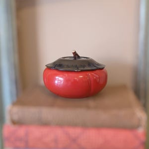 May include: A round, glossy red ceramic lidded container shaped like a fruit. The lid is dark brown with a textured, leaf-like design and a small, central stem protrusion. The base is smooth, vibrant red, and features subtle vertical indentations.