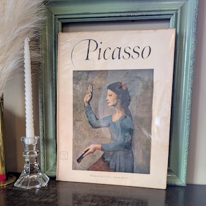 May include: A vintage softcover book titled Picasso featuring a reproduction of a painting of a person in a blue long-sleeved top with one hand raised. The cover includes the text 16 beautiful full color prints at the bottom and a small logo.