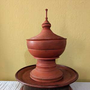 May include: A tiered, reddish-brown wooden decorative object. The top section is a lidded bowl shape, followed by a circular tray, and a base with vertical wooden elements. The object is set against a yellow wall and white cloth.